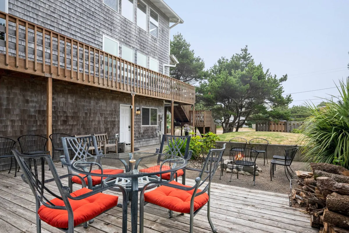 A welcoming outdoor seating area arranged around a stone fire pit on the deck, with the deck railing and natural landscaping visible. The scene captures moments of socializing, outdoor dining, and gathering beside the fire in the evening light.