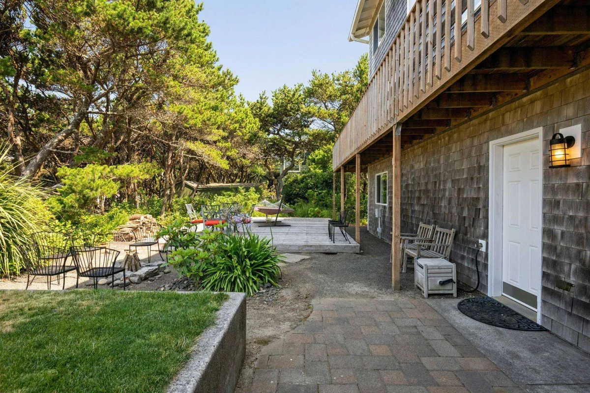 A spacious wooden deck surrounded by lush greenery, featuring a comfortable seating area and a visible pathway leading out to the yard. The tranquil setting suggests a perfect spot for outdoor socializing, relaxing, or gathering around an evening firepit.