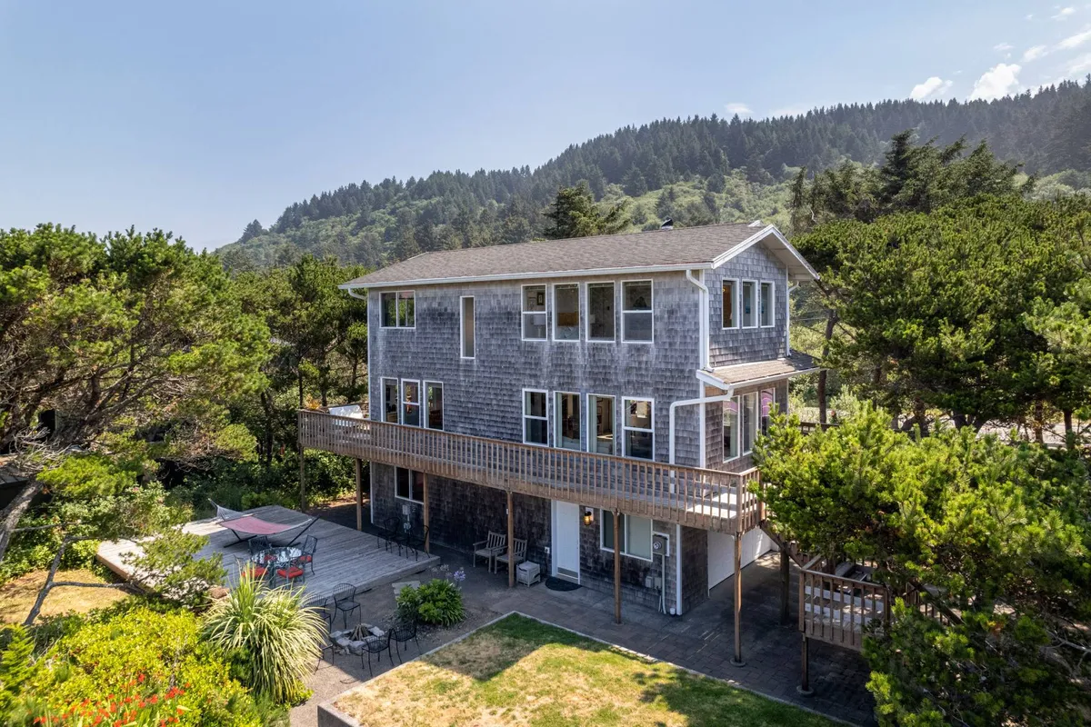 The image features a modern two-story house with expansive windows, a spacious deck outfitted with comfortable seating, and lush greenery surrounding the property. The deck is perfectly positioned for enjoying sunsets and peaceful mornings, with nature and ocean breezes just steps away.