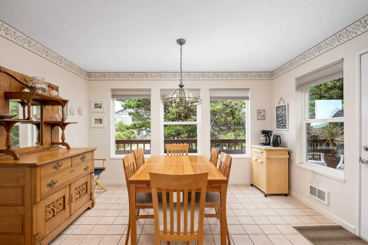 A warm dining area centered around a wooden dining table, with large windows letting in natural light and a wooden sideboard to one side. The setting feels intimate and is clearly designed for family meals or social gatherings.