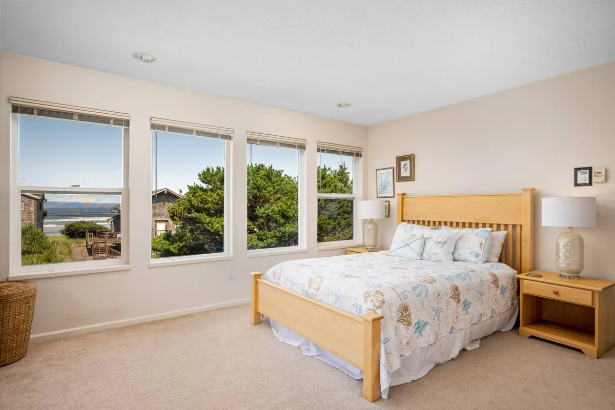 A serene bedroom with a bed made up with decorative pillows, large windows offering views of the outside, and cozy nightstands with lamps. The ambiance is restful and intimate, suggesting a retreat within the home where guests can wake up to tranquil views and soft natural light.