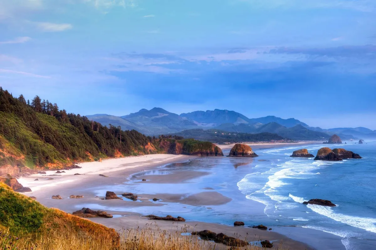 A breathtaking view of the beach and Pacific Ocean, highlighted by striking rock formations, distant mountains, and an expansive sky. The serene scene encapsulates the natural beauty and dramatic coastal landscape of Cape Kiwanda.