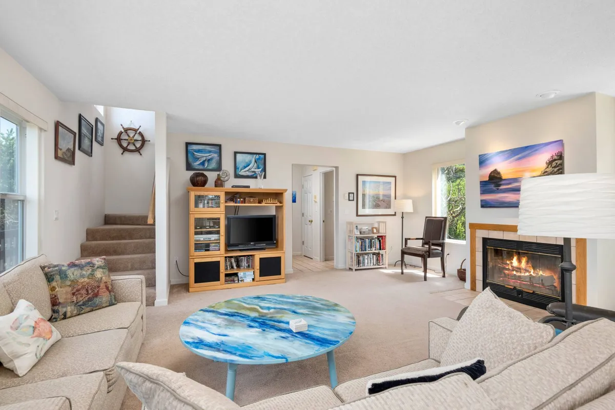 A bright, welcoming living room featuring a colorful round coffee table, a cozy fireplace, art on the walls, and a comfortable sofa. The arrangement invites relaxation and socialization, with thoughtful design touches and warm firelight creating a gathering-friendly space.