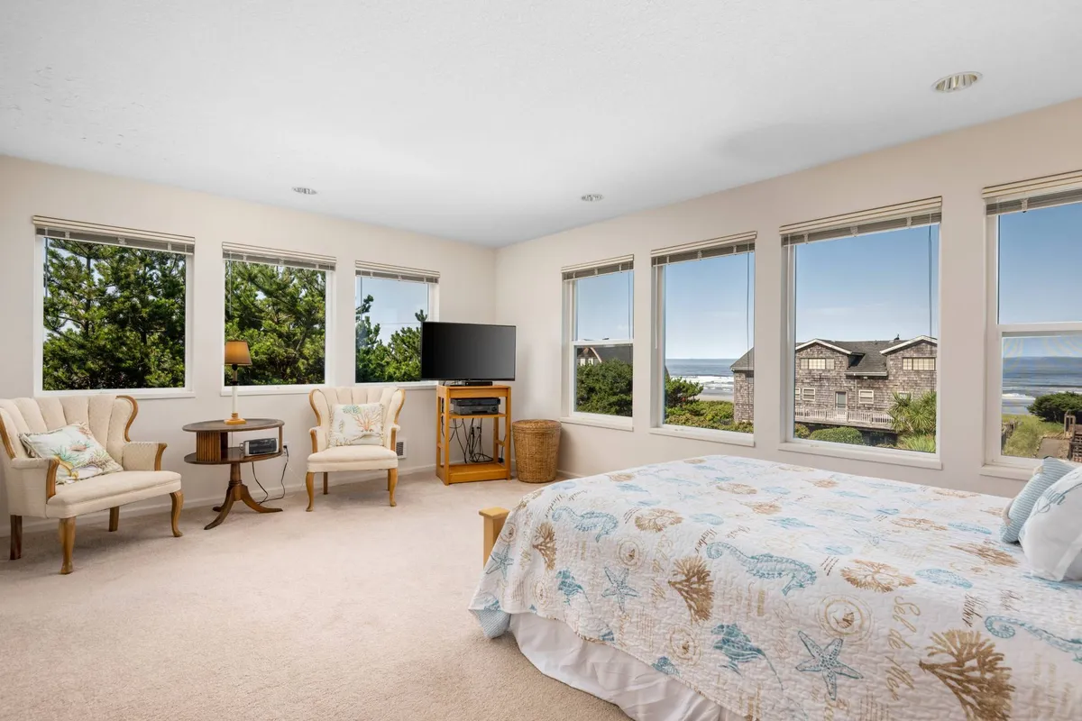 A calm and coastal-inspired bedroom with a sea-themed quilt, large windows offering a panoramic ocean view, and a cozy seating area by the window. The abundant natural light emphasizes the room's relaxing retreat vibe.