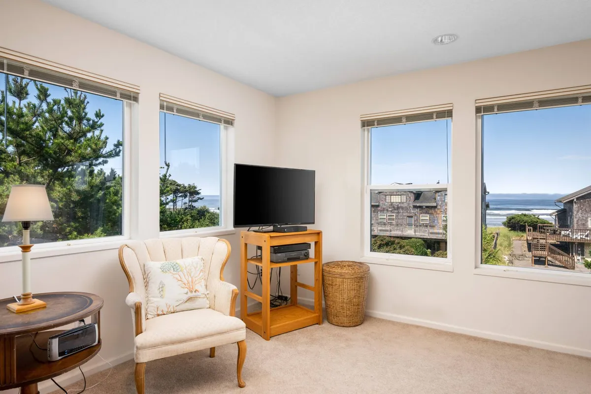A calm, cozy interior with large windows offering scenic views, a comfortable armchair, a television set, a wooden side table, and a wicker basket. Natural light fills the space, which is perfect for watching TV, reading, or simply enjoying time with family.