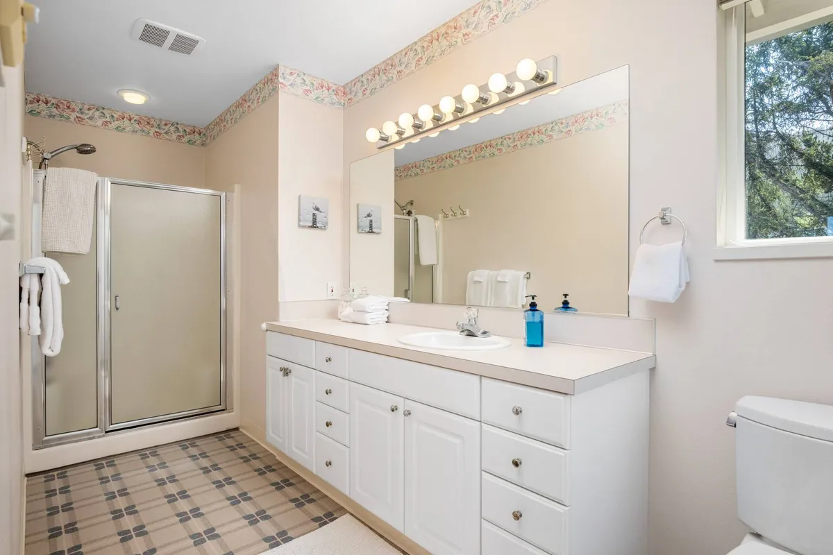 A bright bathroom with a large mirror, vanity, and a shower stall. Morning sunlight pours in through the window, lending a spa-like, refreshing atmosphere perfect for relaxation and self-care.